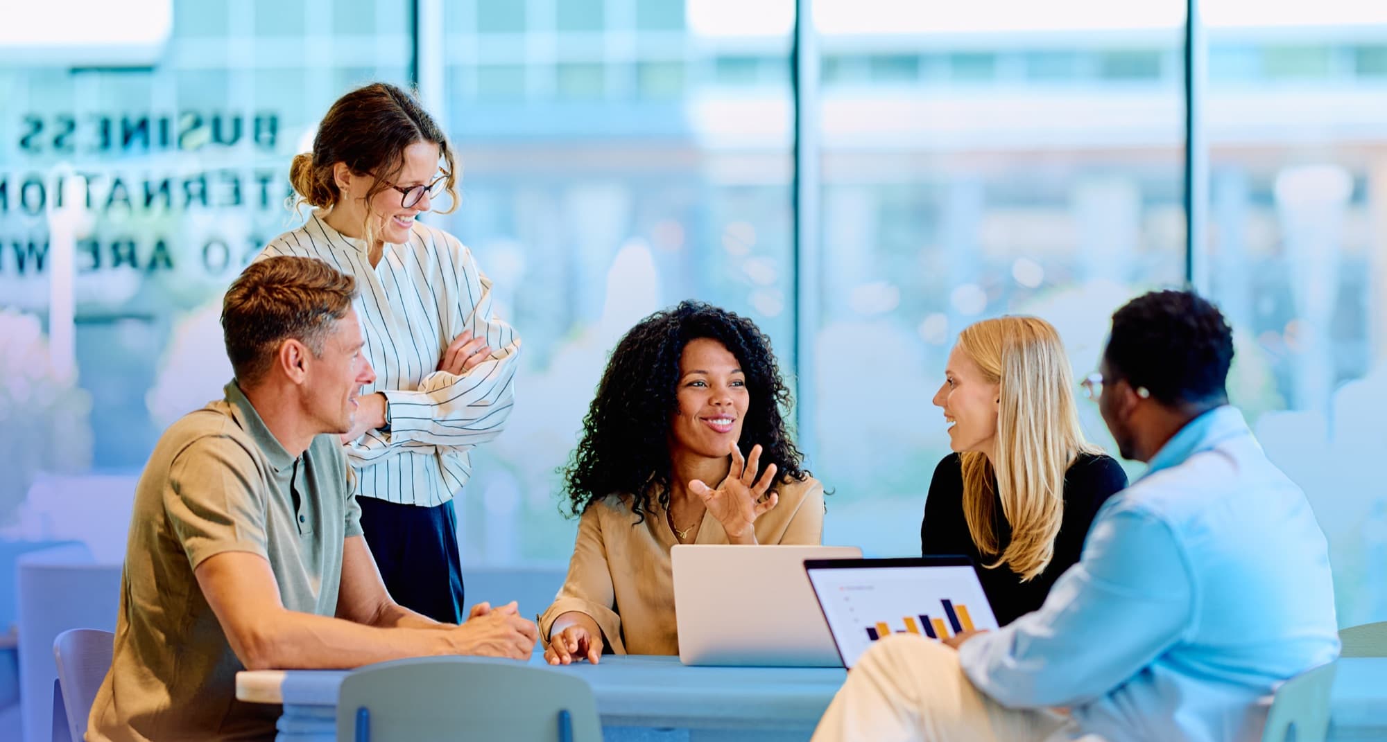 Five Arbitr colleagues gathered around a table in a bright office, smiling and discussing data on a laptop