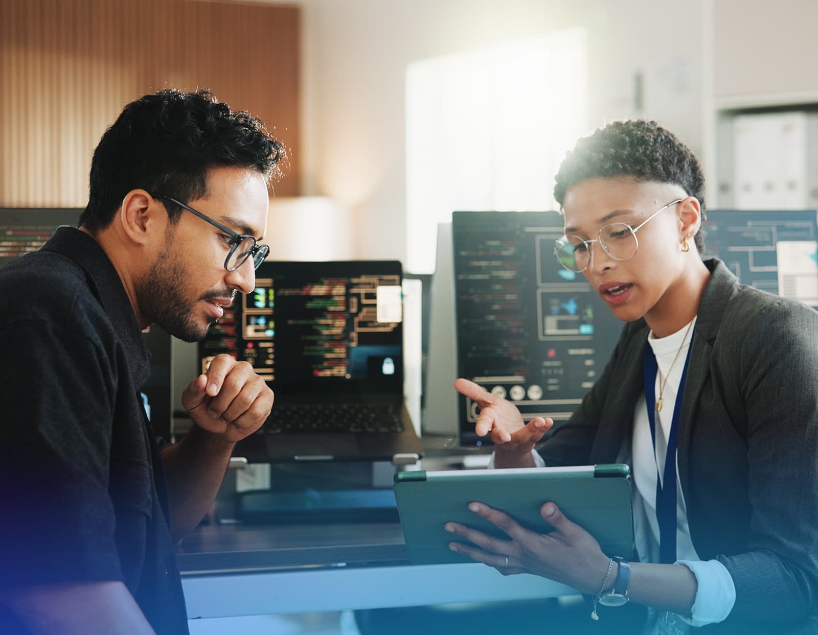 Two colleagues reviewing a tablet together in front of monitors showing code and dashboards